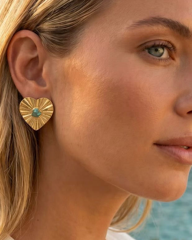 A close-up of a woman wearing a textured gold heart-shaped earring with a small green stone in the center, set against an outdoor background.