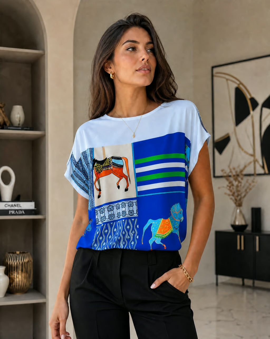 A woman wearing a white short-sleeve t-shirt with a vibrant blue and multicolor patchwork print featuring geometric patterns and stylized camel illustrations, paired with black trousers in a modern indoor setting.