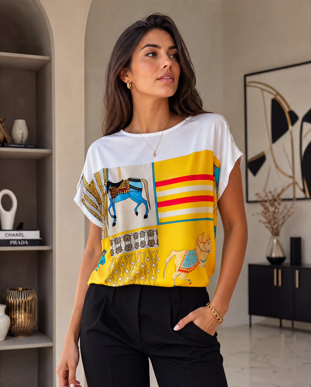 A woman wearing a white short-sleeve top with a vibrant yellow, red, and blue graphic print panel on the front, paired with black trousers, standing in a modern interior setting.