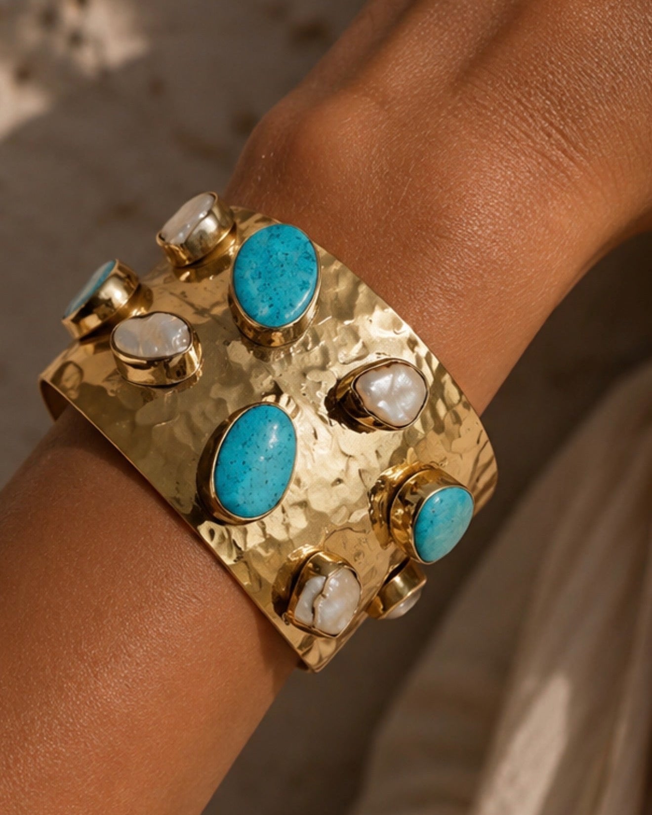 A close-up of a wide gold cuff bracelet worn on a wrist, decorated with several oval turquoise-blue stones and smaller white marbled stones.