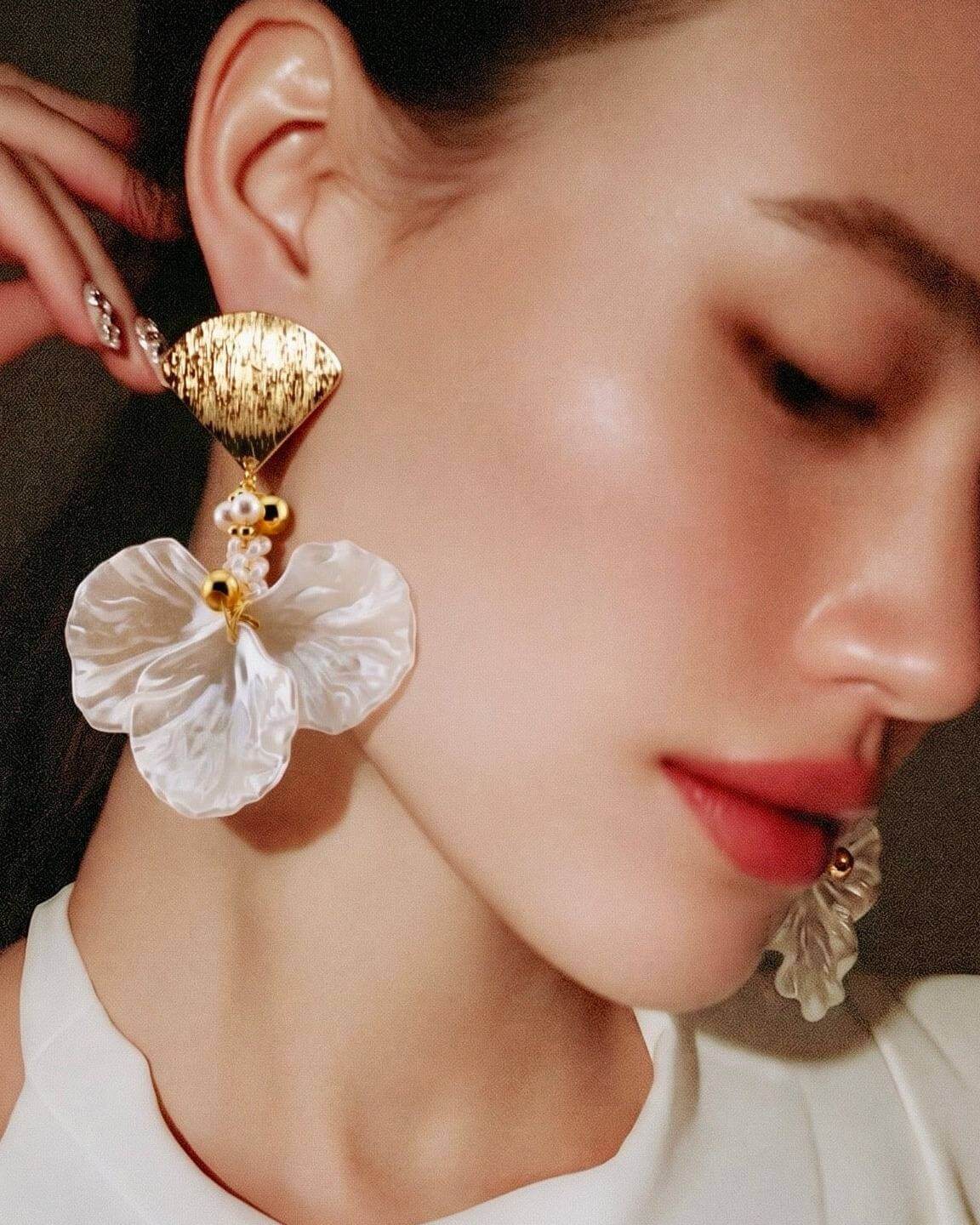 A close-up of a model wearing a large statement earring with a textured gold top, small pearl-like beads, and a translucent white flower dangle.
