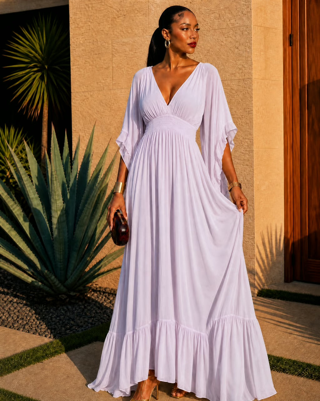 A woman wearing a flowing lavender maxi dress with a deep V-neckline and elbow-length sleeves, standing outdoors in front of a beige stucco wall and agave plant.