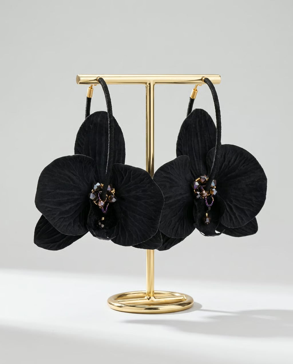 A pair of black orchid-shaped hoop earrings displayed on a gold-tone stand against a light gray studio background.
