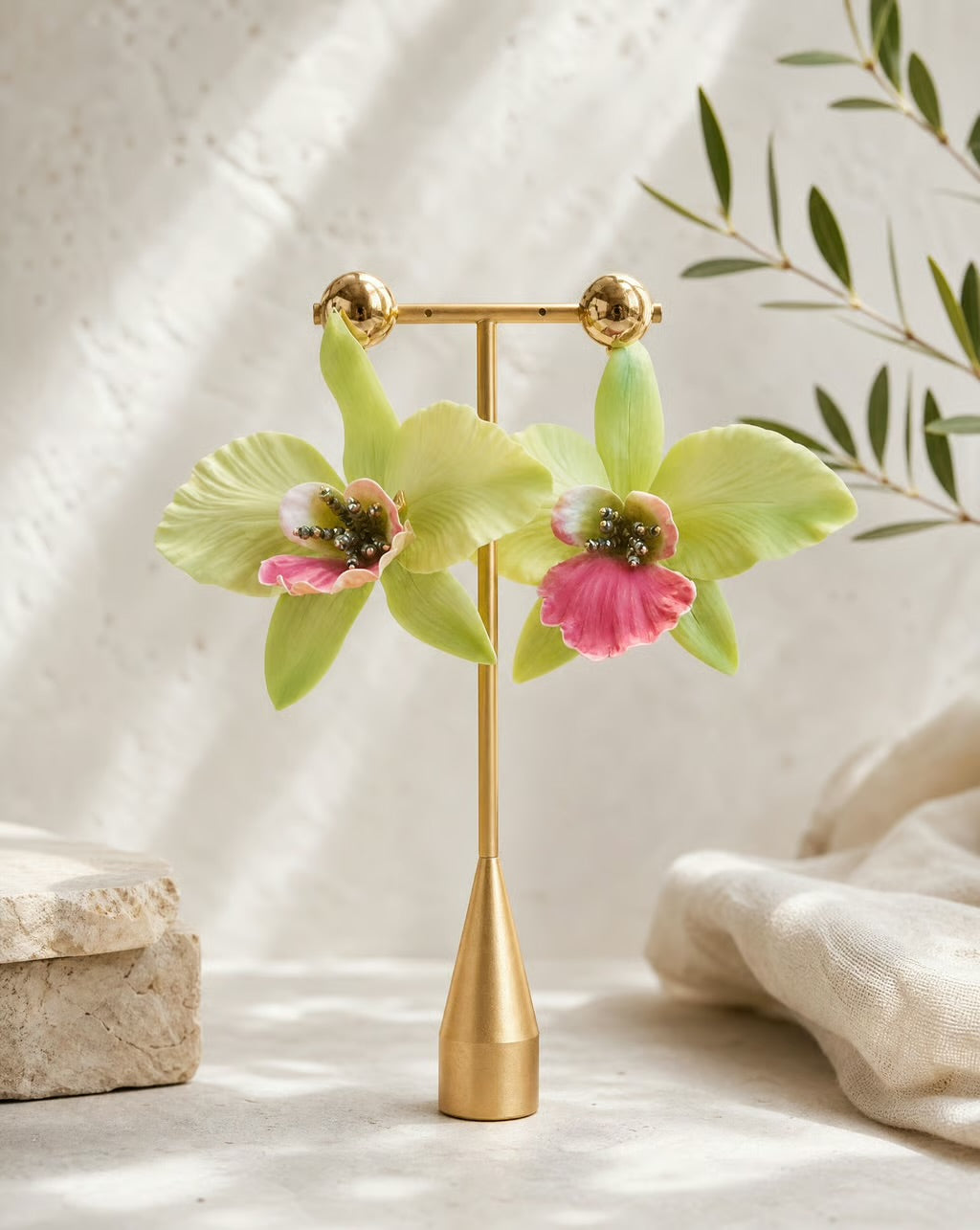 A pair of gold hoop earrings with large light green orchid flower charms and pink centers, displayed on a gold stand against a softly lit neutral background with a linen cloth and leafy branch.
