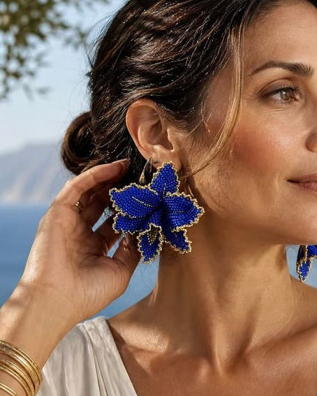 A woman wearing large blue beaded bow earrings with gold trim, shown in a close-up outdoor portrait with flowers and a blurred scenic background.