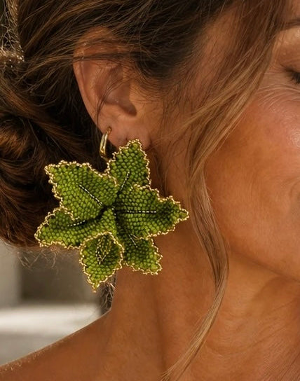 A woman wearing large, textured green grapevine-style drop earrings, shown in a close-up outdoor portrait.