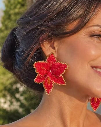 A woman wearing large red flower-shaped earrings with gold trim, photographed outdoors with blurred greenery and pink flowers in the background.