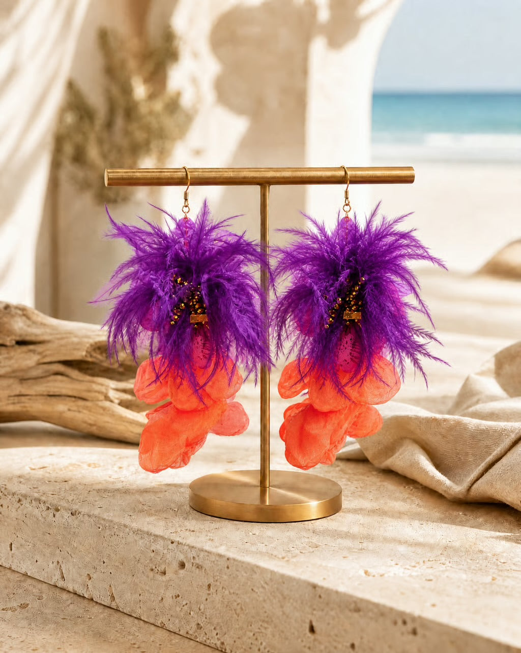 A pair of dangling earrings displayed on a gold stand, featuring fluffy purple feathers transitioning into layered orange petal-like shapes, set against a softly blurred beach and draped fabric background.