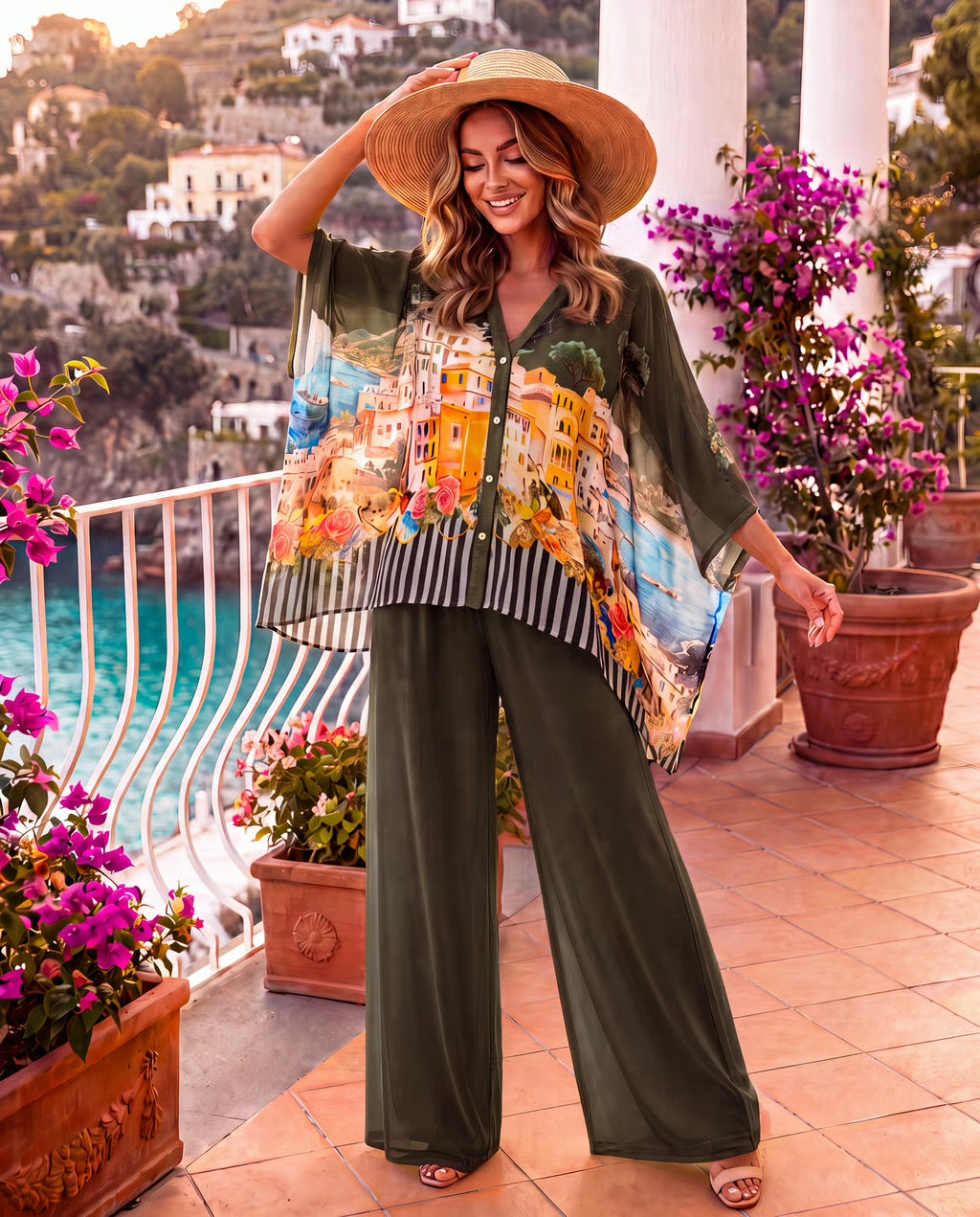 A woman stands on a sunlit balcony wearing a loose olive green two-piece set with a colorful printed cape-style top and wide-leg pants, along with a wide-brim straw hat and sandals. The background shows flowers, terracotta pots, white columns, and a coastal town scene.